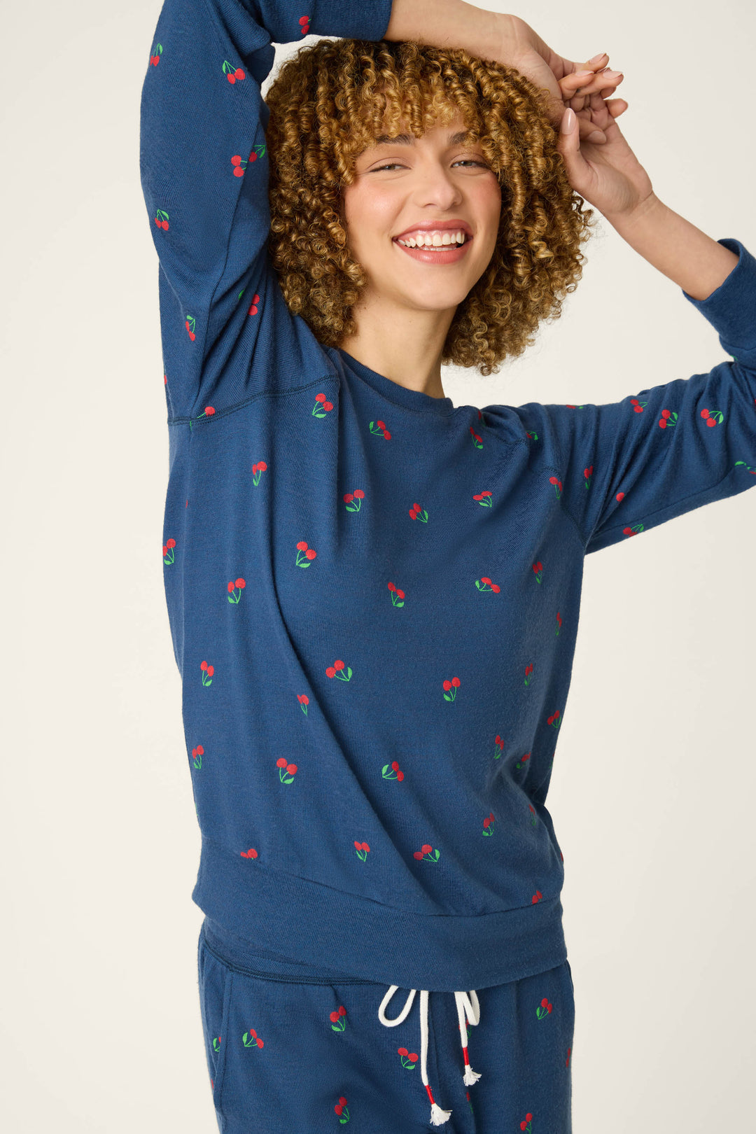 A person with curly hair is smiling and raising one arm while wearing a long sleeve top with a navy background and cherry print.
