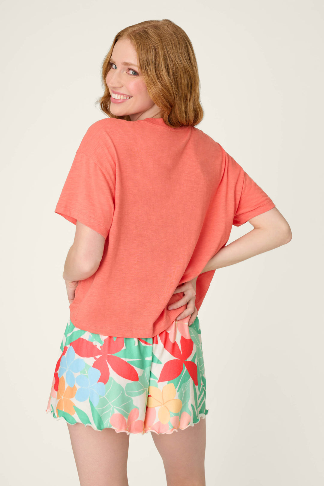 A woman faces away, smiling over her shoulder in a loose graphic tee and green floral shorts, evoking relaxed coastal club vibes.