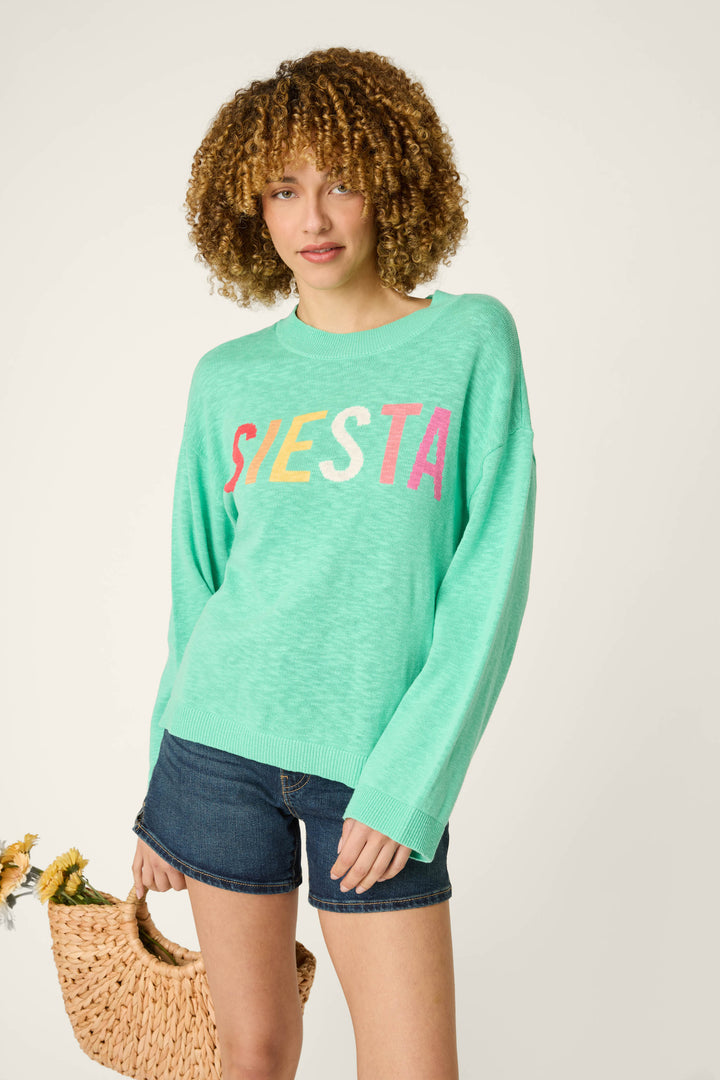 A person stands against a plain background wearing a mint green sweater with colorful lettering, denim shorts, and holding a woven bag of flowers.