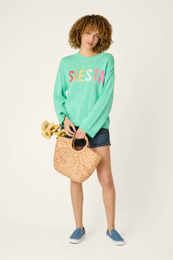A person stands against a plain light background wearing a mint green sweater, denim shorts, blue slip-on shoes, and holding a woven bag of yellow flowers.