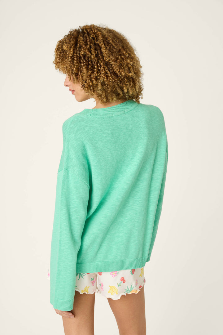 A person with curly hair faces away, wearing a loose, light green sweater and colorful white shorts, set against a plain background.