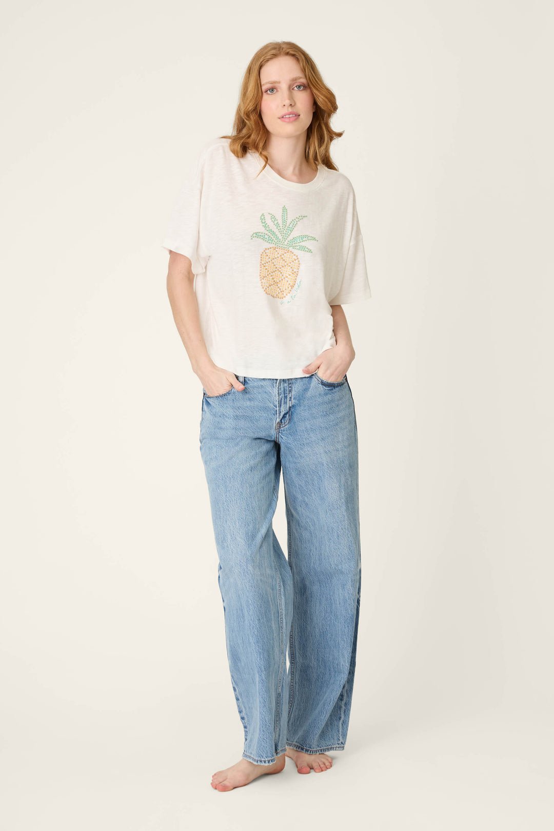 A person stands barefoot against a plain background, hands in pockets, wearing a pineapple-print T-shirt and loose blue jeans.