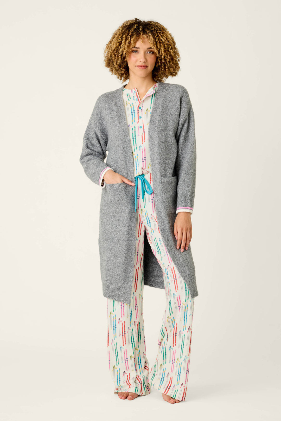 Draped in the P.J. Salvage Après All Day Cardigan Snuggle Sweater, a person stands barefoot against a simple light backdrop, pairing the cozy gray cardigan with white pajama pants and a coordinating top adorned with colorful lines.