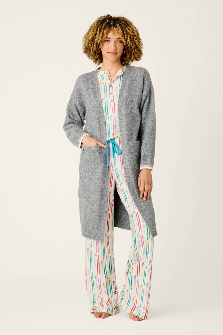Draped in the P.J. Salvage Après All Day Cardigan Snuggle Sweater, a person stands barefoot against a simple light backdrop, pairing the cozy gray cardigan with white pajama pants and a coordinating top adorned with colorful lines.