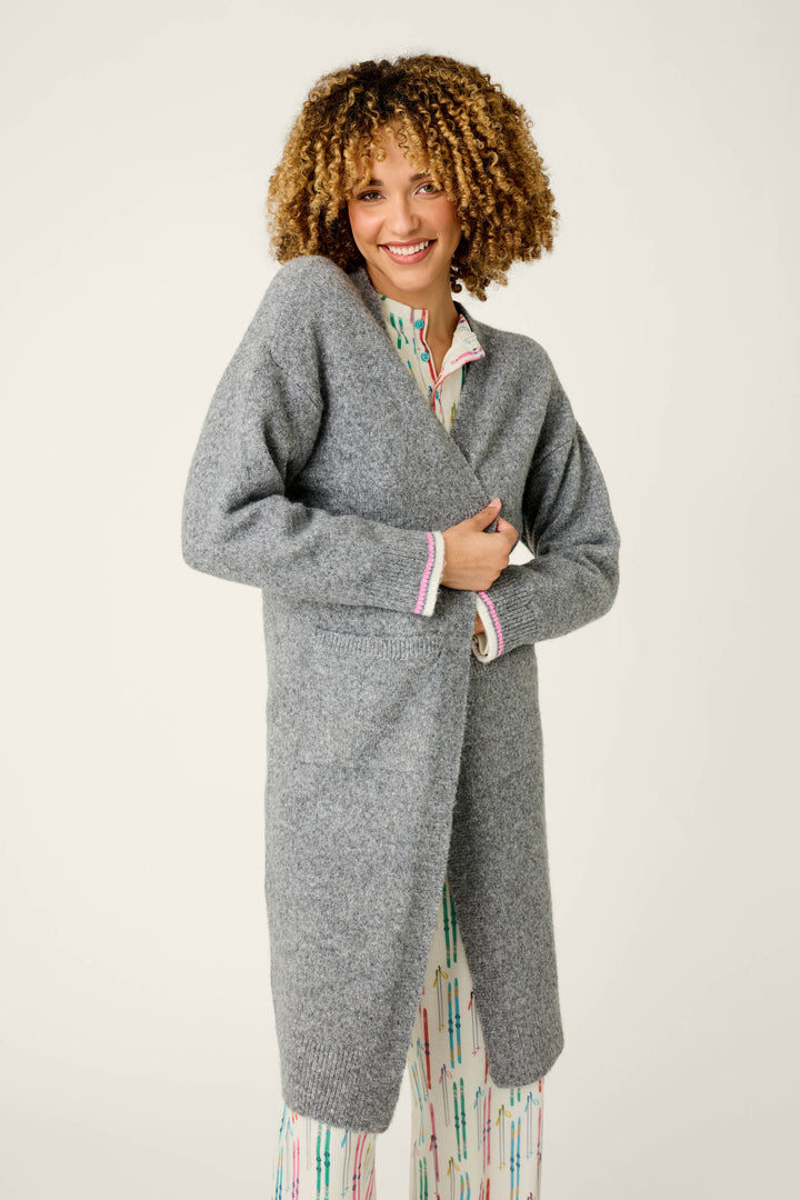 A person with curly hair smiles while wearing the P.J. Salvage Après All Day Cardigan Snuggle Sweater over light-colored pajamas with a colorful pattern, standing against a plain light background.