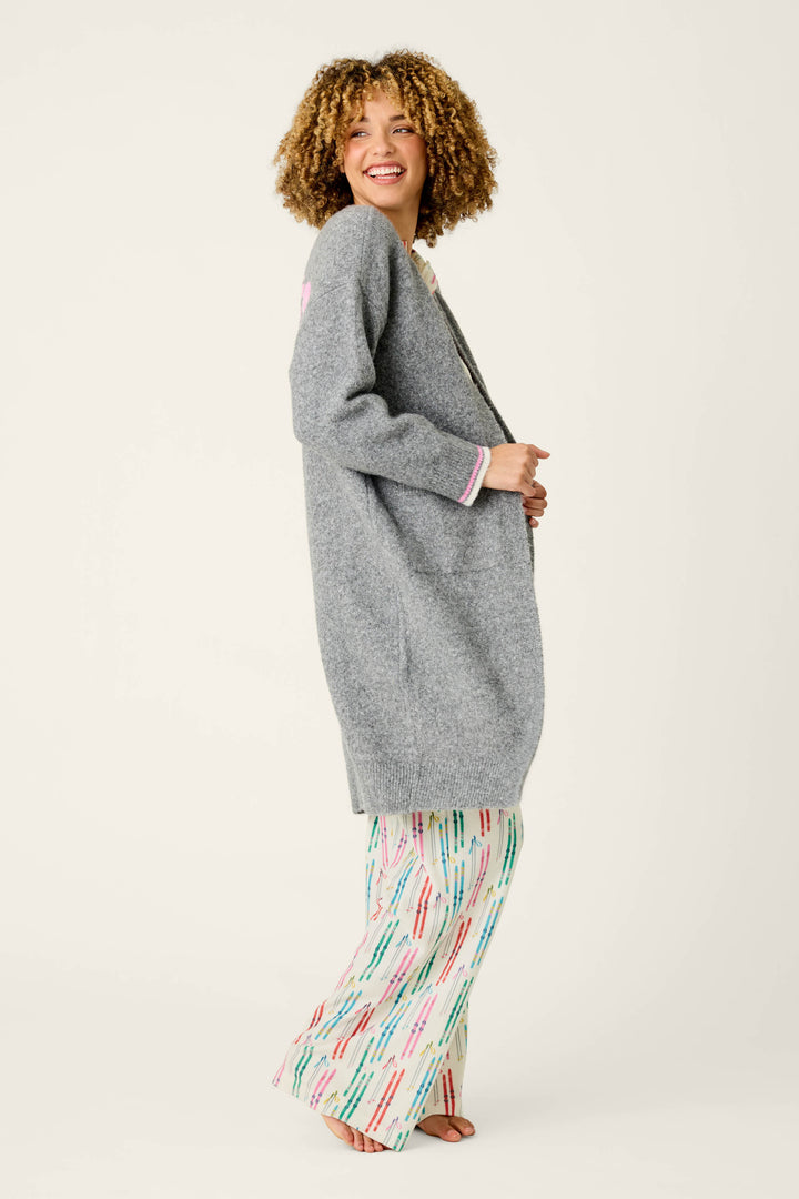 Smiling and looking back, a person stands sideways against a plain light background, wearing the P.J. Salvage Après All Day Cardigan Snuggle Sweater layered over colorful, patterned pajama pants.