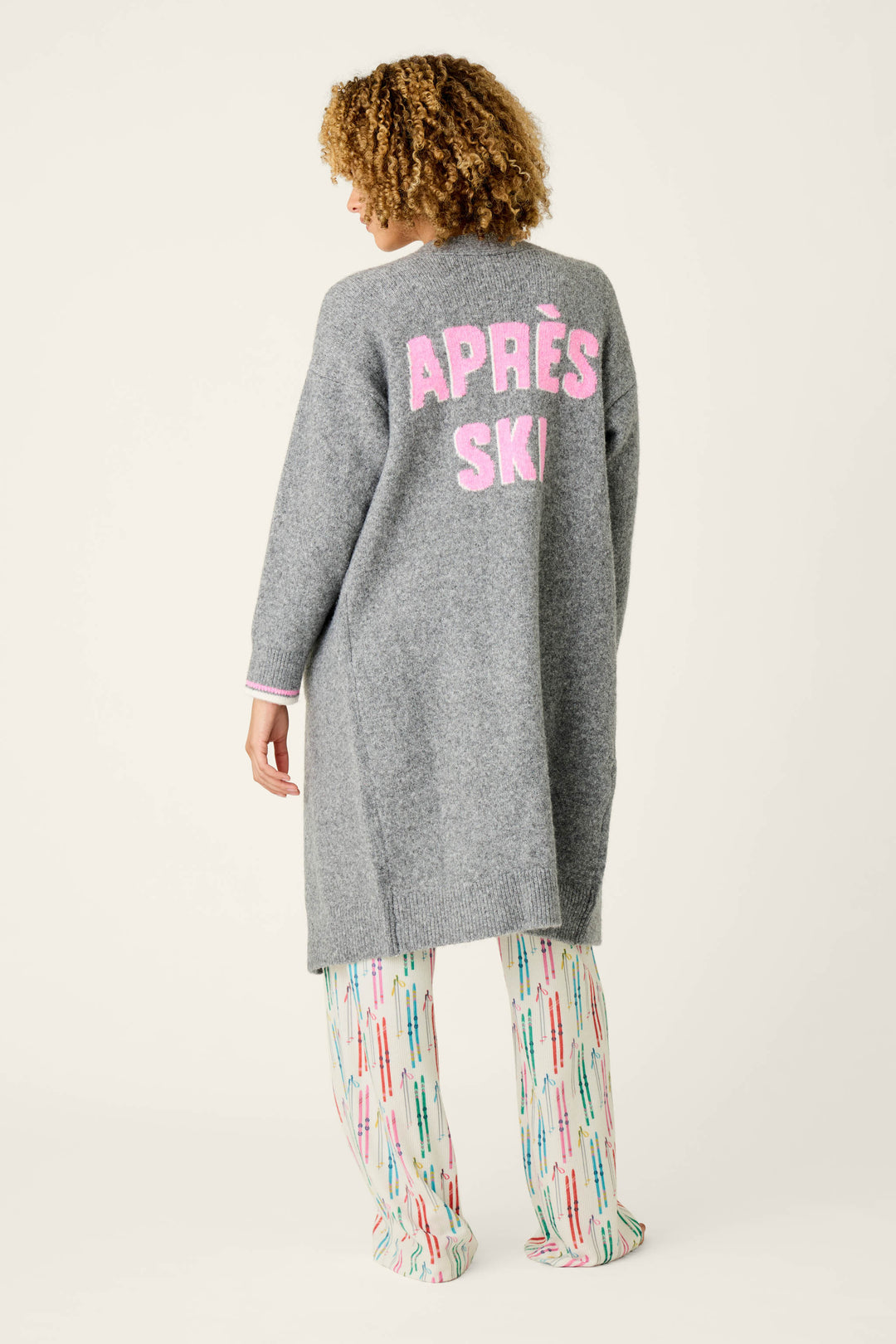 A person with curly hair models the Après All Day Cardigan Snuggle Sweater by P.J. Salvage, a long gray sweater featuring “APRÈS SKI” in pink text on the back, paired with colorful patterned pants and posed against a plain light background.