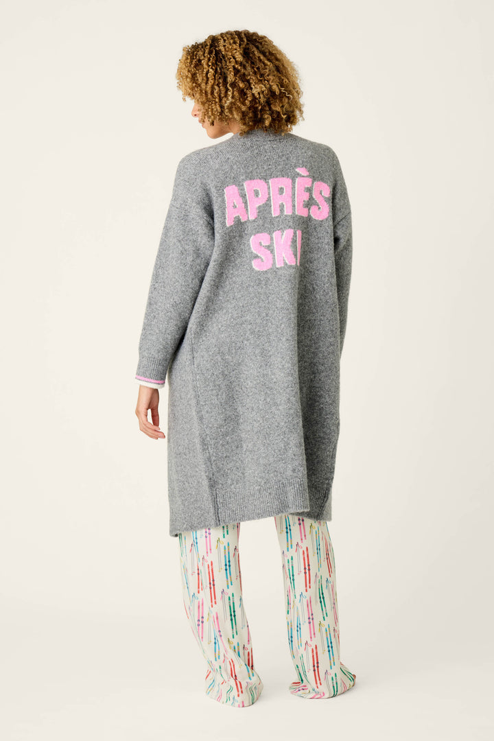 A person with curly hair models the Après All Day Cardigan Snuggle Sweater by P.J. Salvage, a long gray sweater featuring “APRÈS SKI” in pink text on the back, paired with colorful patterned pants and posed against a plain light background.