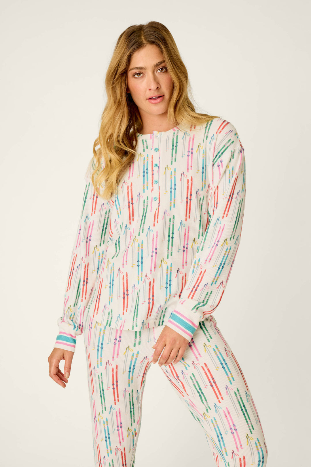 A person with long blonde hair is wearing the P.J. Salvage Après All Day Ski Print Long Sleeve Henley Top, which features a colorful ski-themed pattern on a white background, while standing against a plain light backdrop.