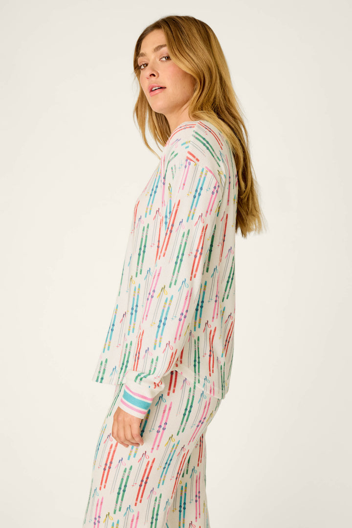 A person with long blonde hair wears the P.J. Salvage Après All Day Ski Print Long Sleeve Henley Top, featuring colorful vertical line patterns, standing sideways against a plain light background.