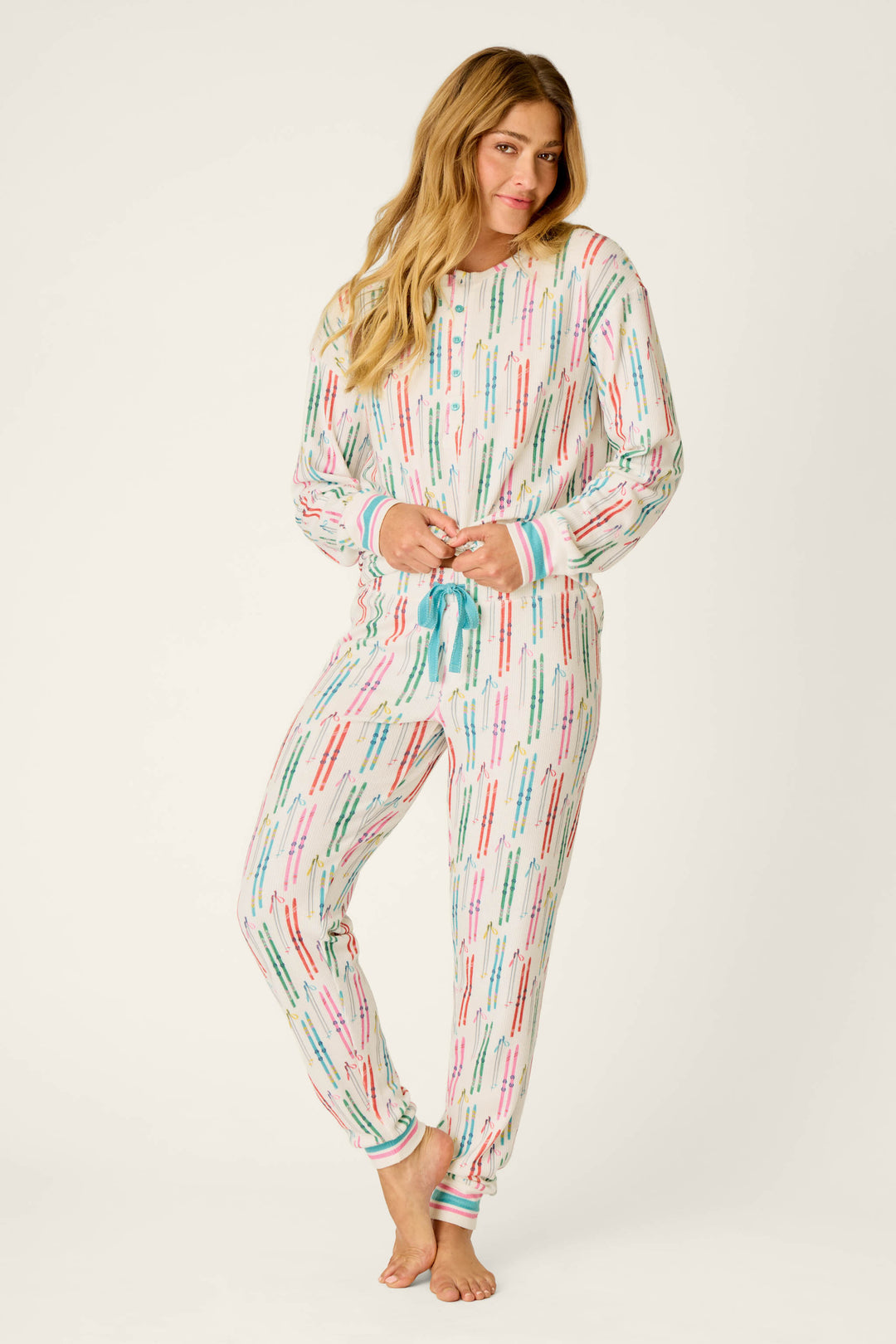 A woman stands barefoot on a plain background with a relaxed expression, holding the waistband of her pajama bottoms while wearing the P.J. Salvage Après All Day Ski Print Long Sleeve Henley Top.