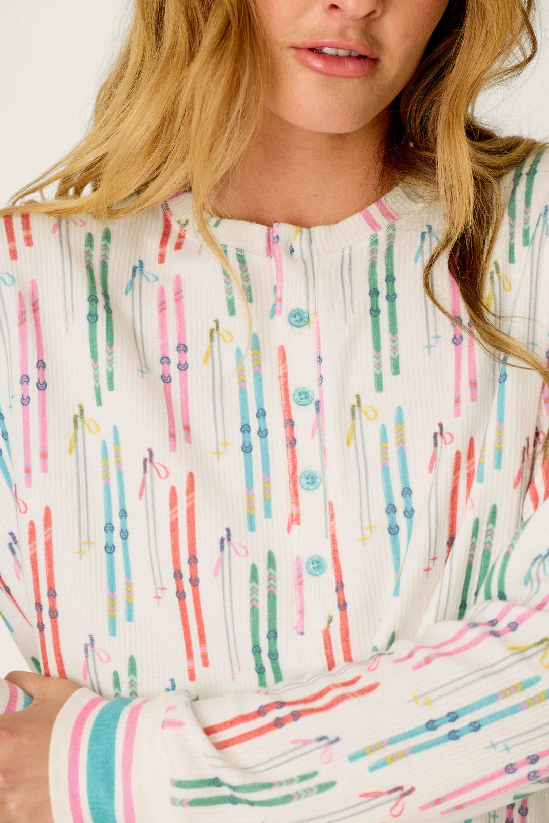 A woman models the P.J. Salvage Après All Day Ski Print Long Sleeve Henley Top, featuring colorful ski and ski pole patterns, turquoise buttons, and pink striped cuffs; image shown from shoulders to mouth.