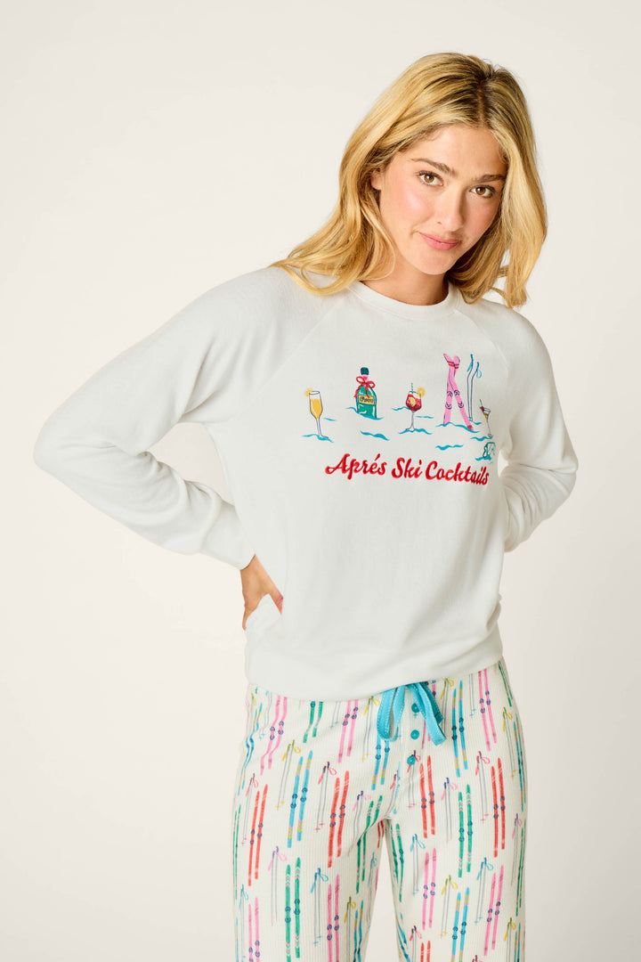 A woman with blonde hair wears the P.J. Salvage Après All Day Après Ski Cocktails Long Sleeve Top, featuring colorful cocktail illustrations and the text “Après Ski Cocktails,” paired with white pajama pants decorated with a ski pole pattern.