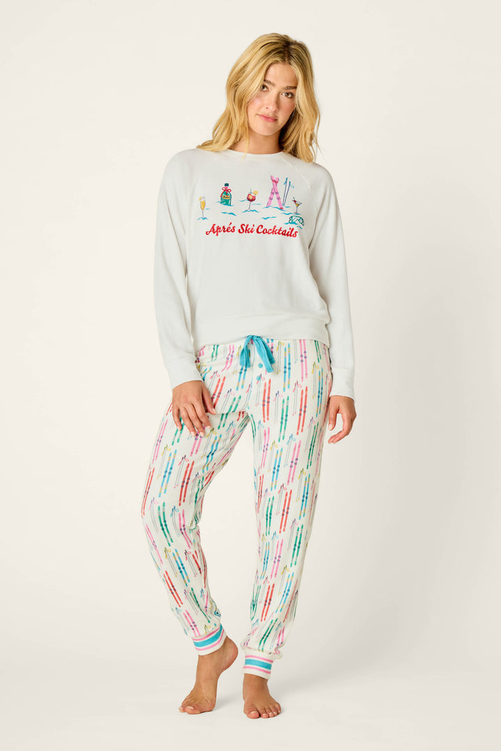A woman with long blonde hair wears the P.J. Salvage Après All Day Après Ski Cocktails Long Sleeve Top—a white sweater with colorful ski-themed designs and “Après Ski Cocktails” text—paired with white pajama pants featuring a multicolored ski pole pattern. She stands barefoot against a plain background.