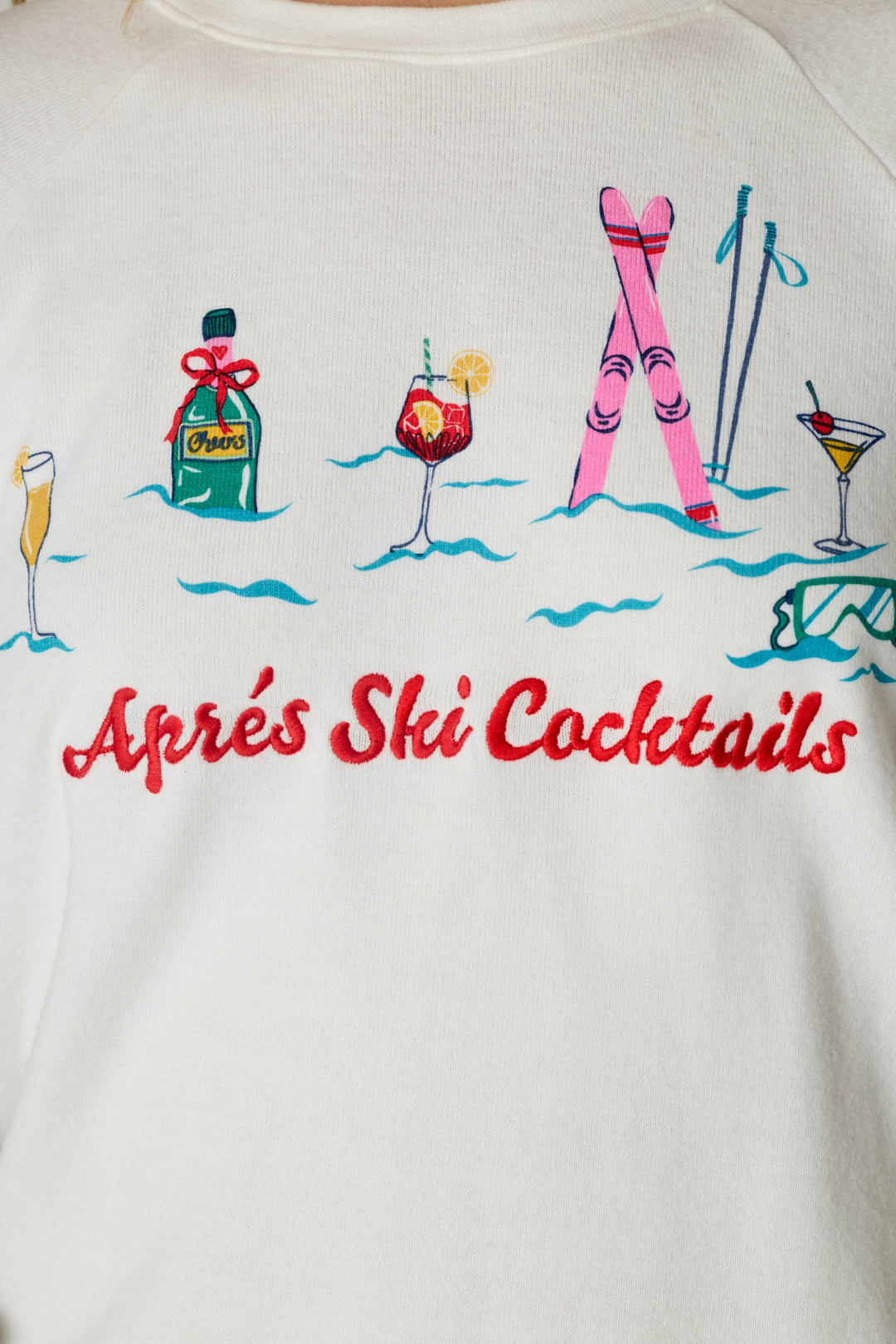 The Après All Day Après Ski Cocktails Long Sleeve Top by P.J. Salvage features colorful embroidery of a bottle, cocktail glasses, pink skis, ski poles, and goggles on a white background, with “Après Ski Cocktails” written in red script beneath the design.