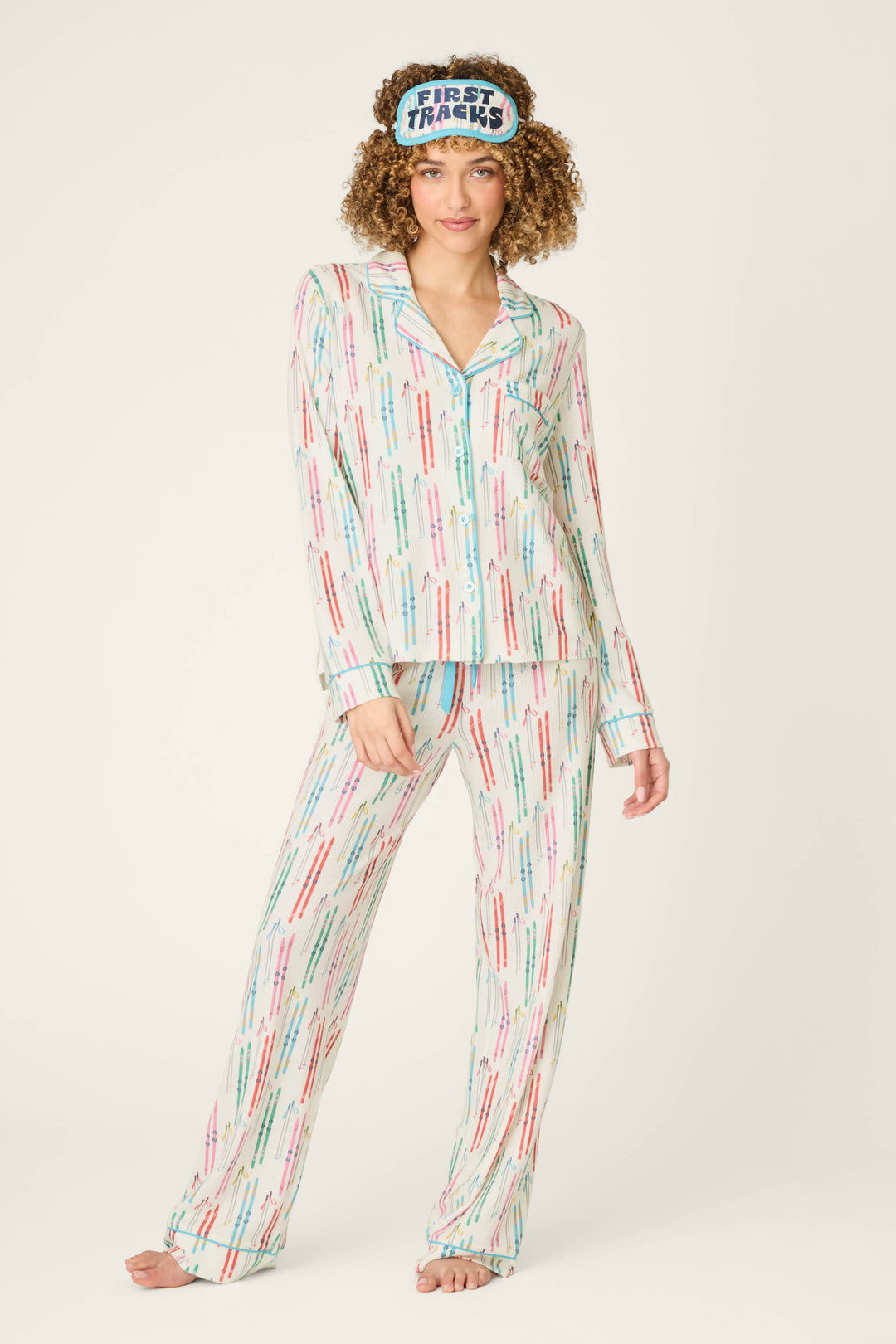 A person is wearing the P.J. Salvage Après All Day Ski Print 3-Piece PJ Set, which features a colorful, repeating text design and comes with a matching sleep mask that says “THANKS THANKS THANKS.” The individual is barefoot and standing against a plain background.