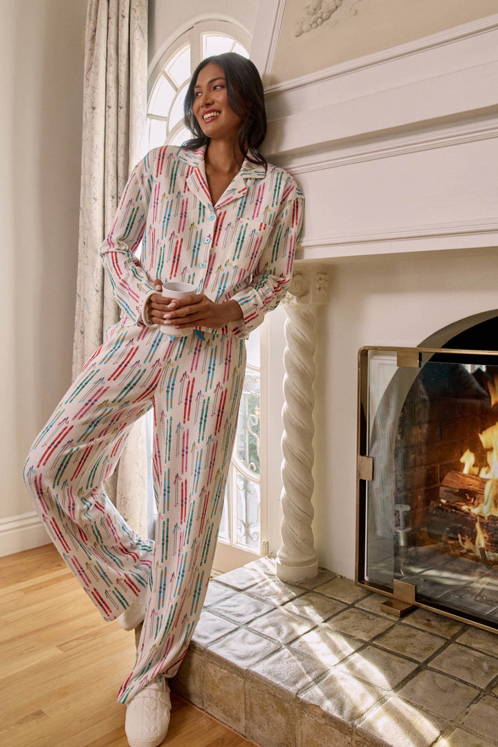 Wearing the Après All Day Ski Print 3-Piece PJ Set by P.J. Salvage, a woman stands smiling by a cozy fireplace in an elegant, sunlit room with wooden floors and a window.