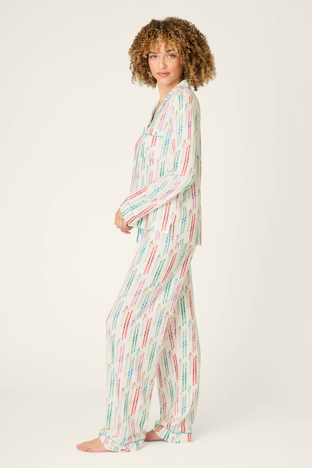 A person stands barefoot, modeling the P.J. Salvage Après All Day Ski Print 3-Piece PJ Set, featuring a playful ski-inspired pattern in pastel hues, viewed from the side against a plain light background.