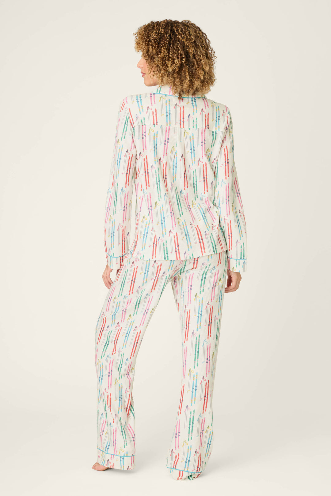 A person stands barefoot, facing away from the camera, wearing the P.J. Salvage Après All Day Ski Print 3-Piece PJ Set—a coordinated long-sleeve pajama set featuring a playful ski-themed print on a white background. The backdrop is plain and light-colored.