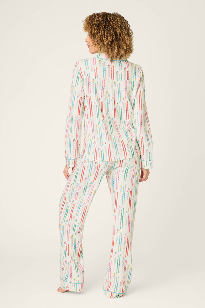 A person stands barefoot, facing away from the camera, wearing the P.J. Salvage Après All Day Ski Print 3-Piece PJ Set—a coordinated long-sleeve pajama set featuring a playful ski-themed print on a white background. The backdrop is plain and light-colored.
