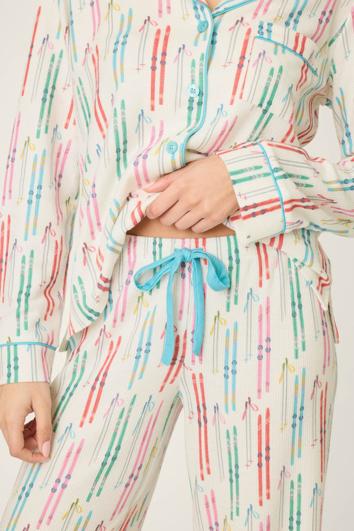 A person is seen wearing the P.J. Salvage Après All Day Ski Print 3-Piece PJ Set, featuring white pajamas adorned with colorful ski pole patterns and blue piping. The pajama pants include a blue drawstring, and only the torso and part of the left hand are visible.