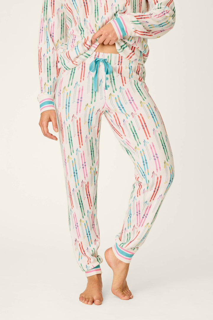 A person wearing P.J. Salvage Après All Day Ski Print Jammie Pants—colorful, patterned pajama pants with a blue drawstring—paired with a matching long-sleeve top, standing barefoot against a plain background.