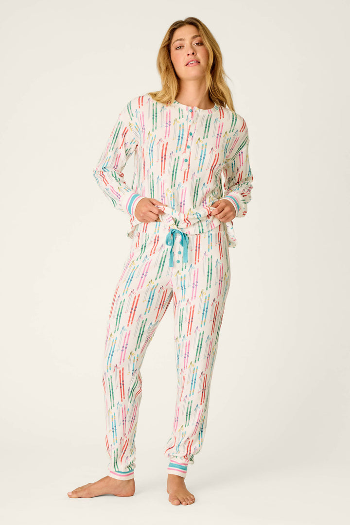 A woman stands barefoot wearing the Après All Day Ski Print Jammie Pants by P.J. Salvage, featuring a playful print of colorful pen or marker-like patterns on a white background. The pajama pants have long legs and blue drawstrings at the waist, paired with a matching long-sleeve top.