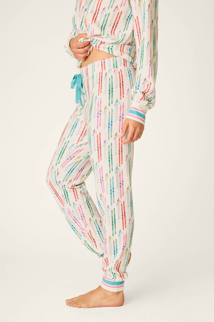 A person stands barefoot against a plain background, wearing P.J. Salvage Après All Day Ski Print Jammie Pants with a matching long-sleeve top featuring colorful crayon patterns. The relaxed-fit outfit includes jogger-style pants with a drawstring waistband.