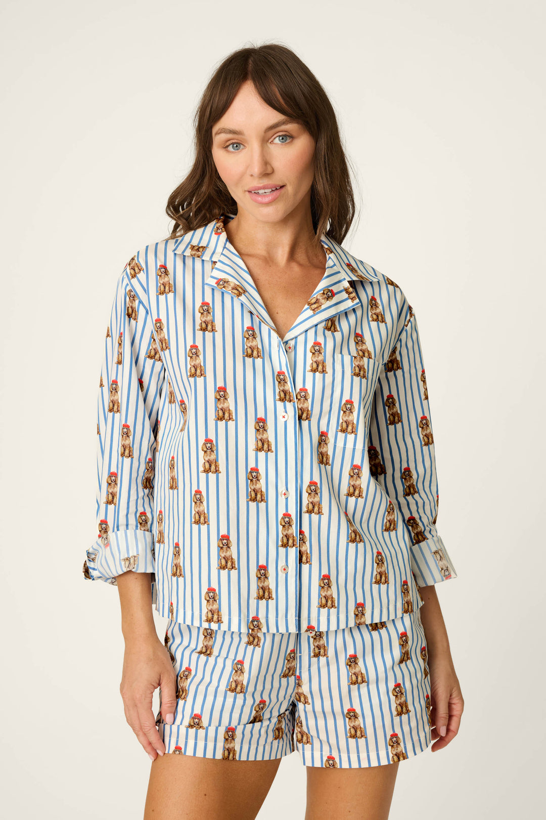A woman wears the P.J. Salvage Afterparty Poplin Stripe & Poodle Print Long Sleeve Top, featuring blue stripes and small brown poodle prints, while standing against a plain light background and looking at the camera with a slight smile.