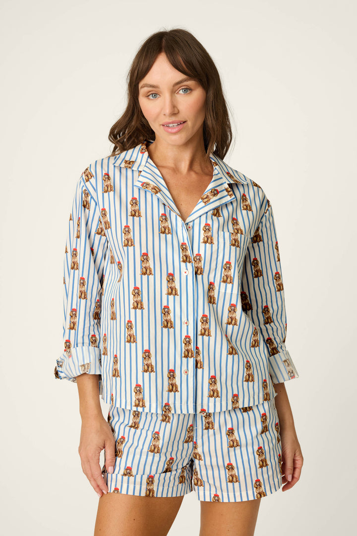 A woman wears the P.J. Salvage Afterparty Poplin Stripe & Poodle Print Long Sleeve Top, featuring blue stripes and small brown poodle prints, while standing against a plain light background and looking at the camera with a slight smile.
