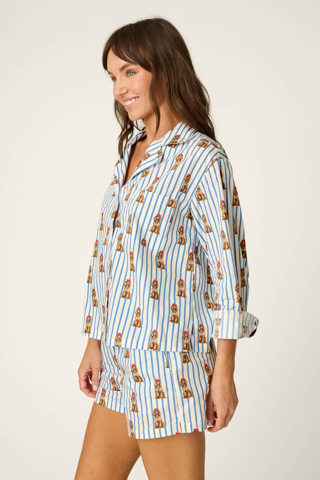 A woman wearing the P.J. Salvage Afterparty Poplin Stripe & Poodle Print Long Sleeve Top stands sideways and smiles against a plain light background.