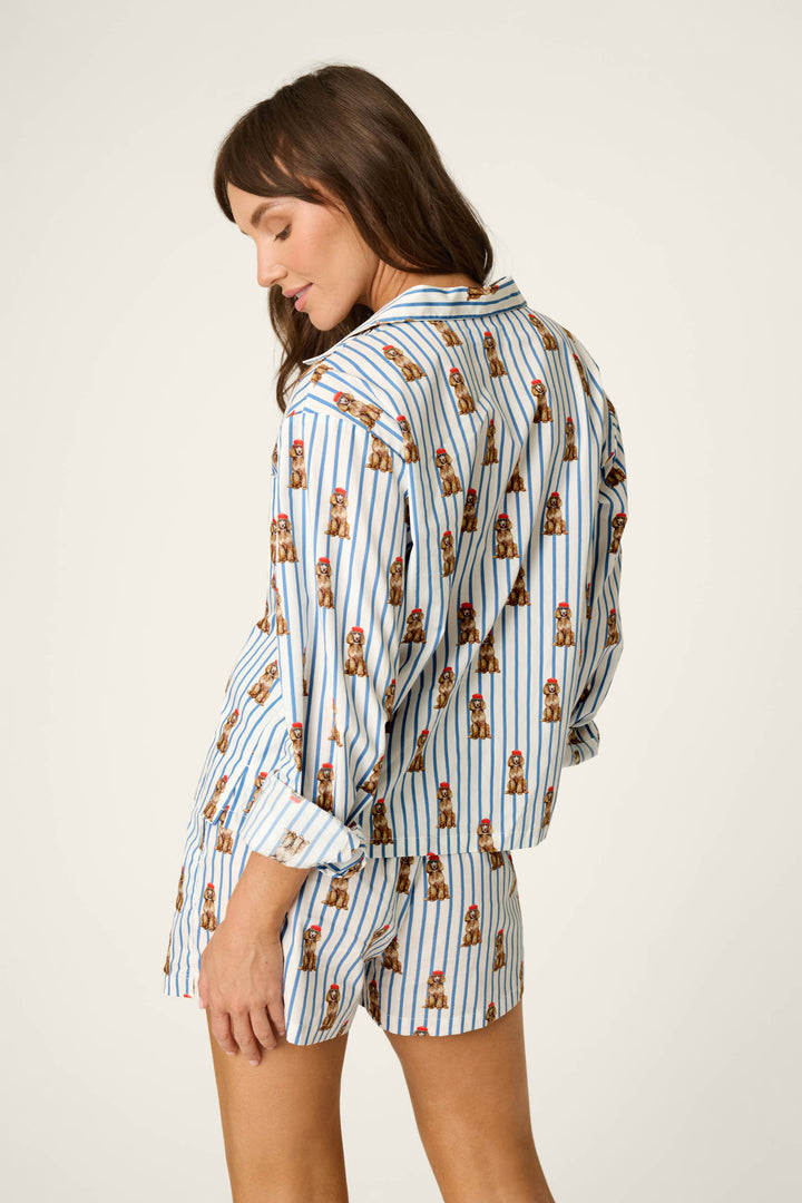 A person stands looking over their shoulder, wearing the P.J. Salvage Afterparty Poplin Stripe & Poodle Print Long Sleeve Top, a matching pajama set featuring blue and white stripes with a poodle print. The background is plain and light colored.