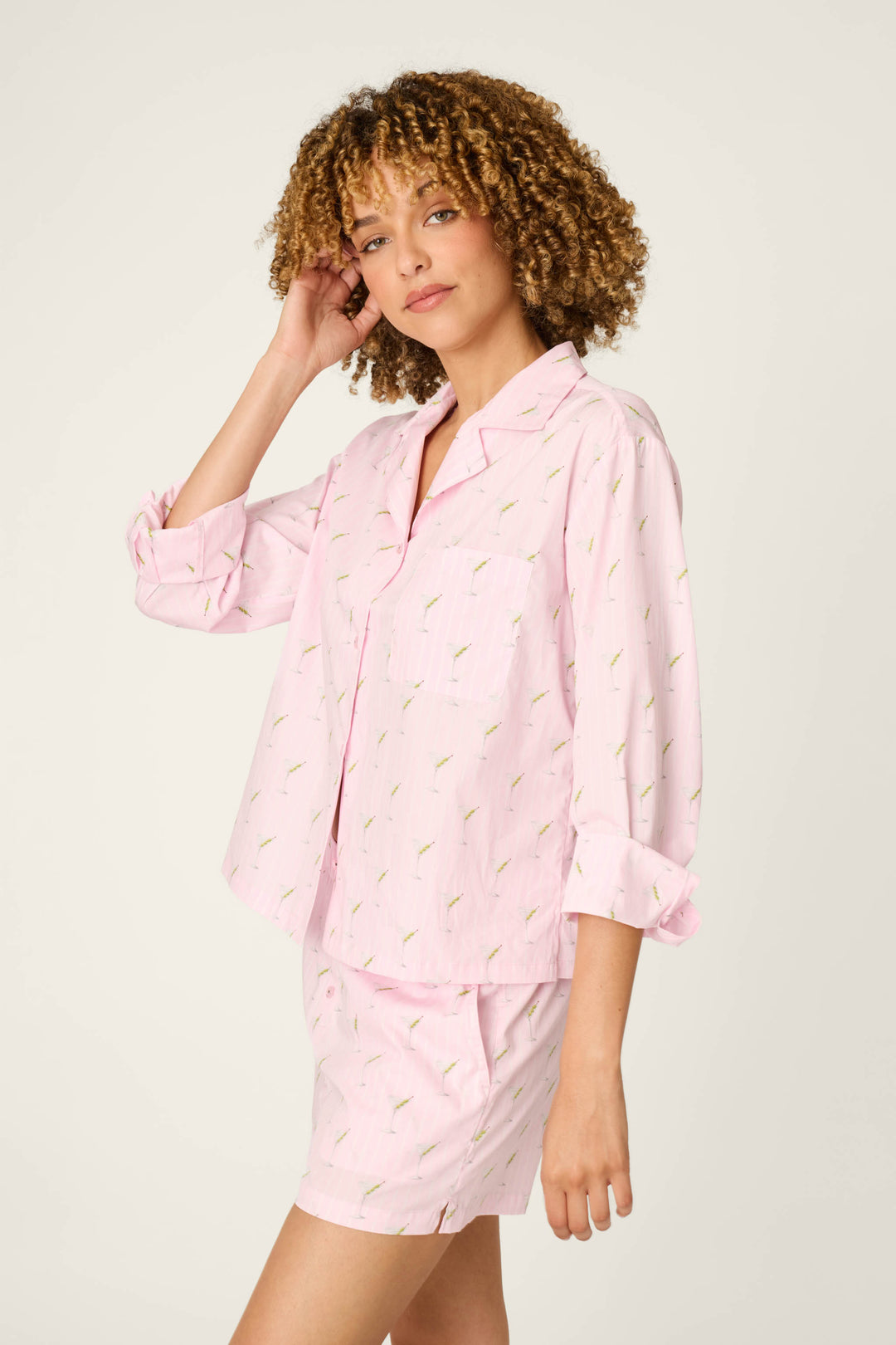 A woman with curly hair models the P.J. Salvage Afterparty Poplin Stripe & Martini Print Long Sleeve Top. She stands against a plain background, facing slightly to the side and touching her hair with one hand.