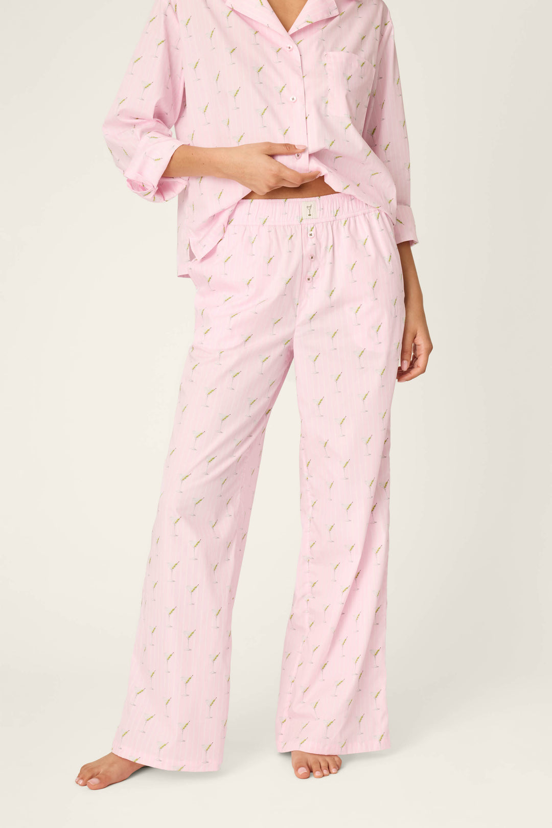 A person is standing barefoot on a neutral background, wearing P.J. Salvage Afterparty Poplin Stripe & Martini Print Pajama Pants with a coordinating light pink, long-sleeve top featuring a small print pattern, and lifting the top slightly to reveal the waistband.