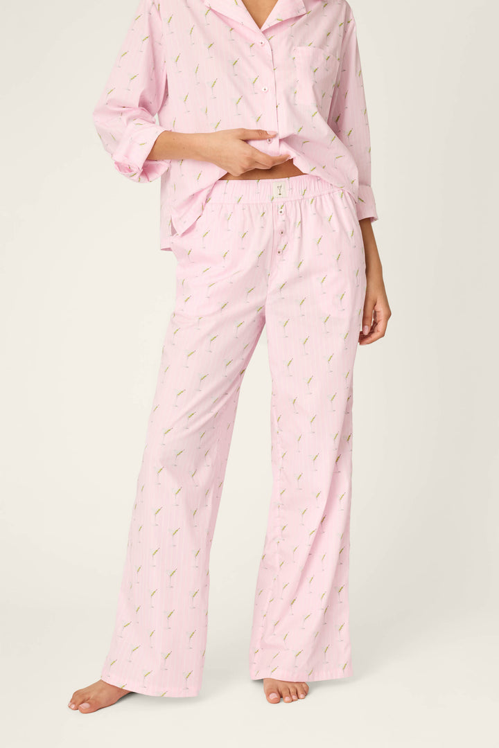A person is standing barefoot on a neutral background, wearing P.J. Salvage Afterparty Poplin Stripe & Martini Print Pajama Pants with a coordinating light pink, long-sleeve top featuring a small print pattern, and lifting the top slightly to reveal the waistband.