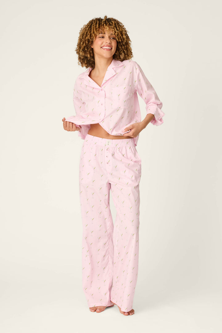 A woman with curly hair smiles, standing barefoot against a plain light background. She lifts her pajama top slightly to reveal the Afterparty Poplin Stripe & Martini Print Pajama Pants by P.J. Salvage, featuring a subtle pink pattern.