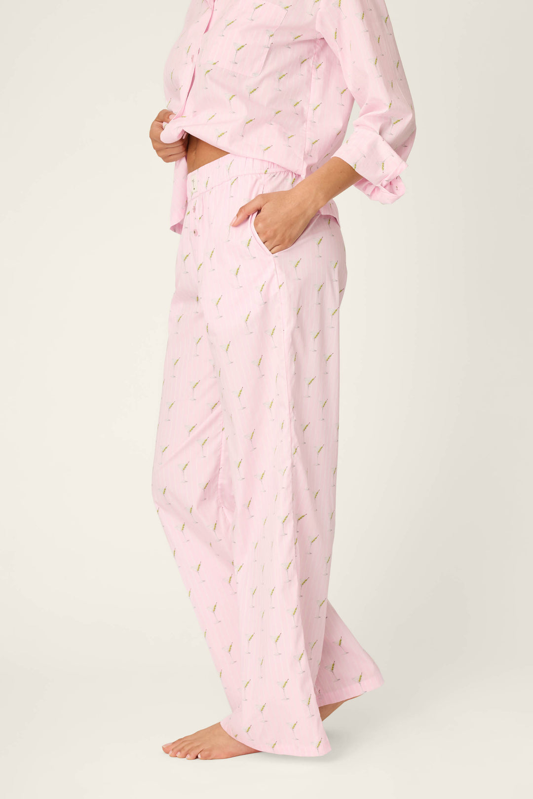 A person wearing P.J. Salvage Afterparty Poplin Stripe & Martini Print Pajama Pants stands barefoot on a light background, with one hand in a pocket and the other holding up the front of the pajama top.