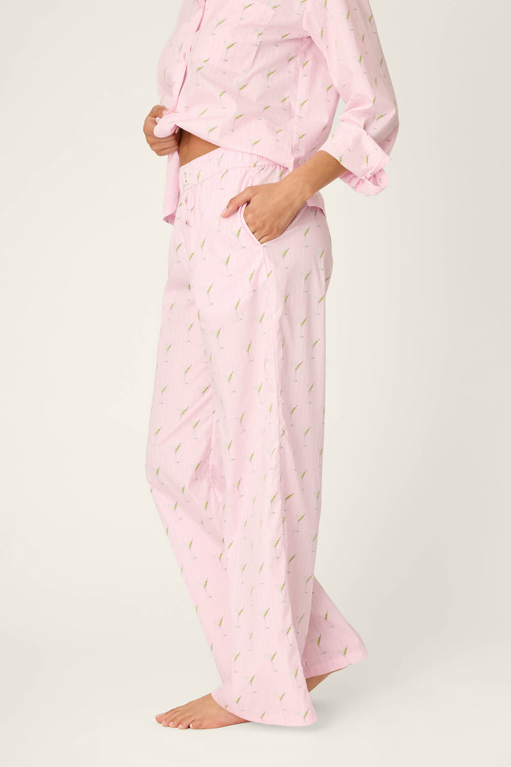 A person wearing P.J. Salvage Afterparty Poplin Stripe & Martini Print Pajama Pants stands barefoot on a light background, with one hand in a pocket and the other holding up the front of the pajama top.
