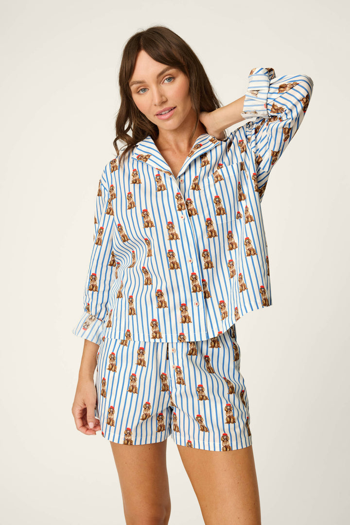 A woman with brown hair poses against a plain beige background wearing the P.J. Salvage Afterparty Poplin Stripe & Poodle Print Long Sleeve & Shorts PJ Set, which features blue and white stripes with a playful brown poodle print. The set includes a long-sleeve top and matching shorts.