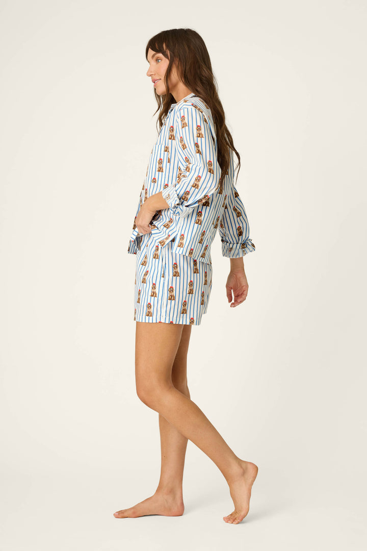 A woman stands barefoot in profile against a plain light background, wearing the P.J. Salvage Afterparty Poplin Stripe & Poodle Print Long Sleeve & Shorts PJ Set—featuring blue and white vertical stripes with playful poodle prints.