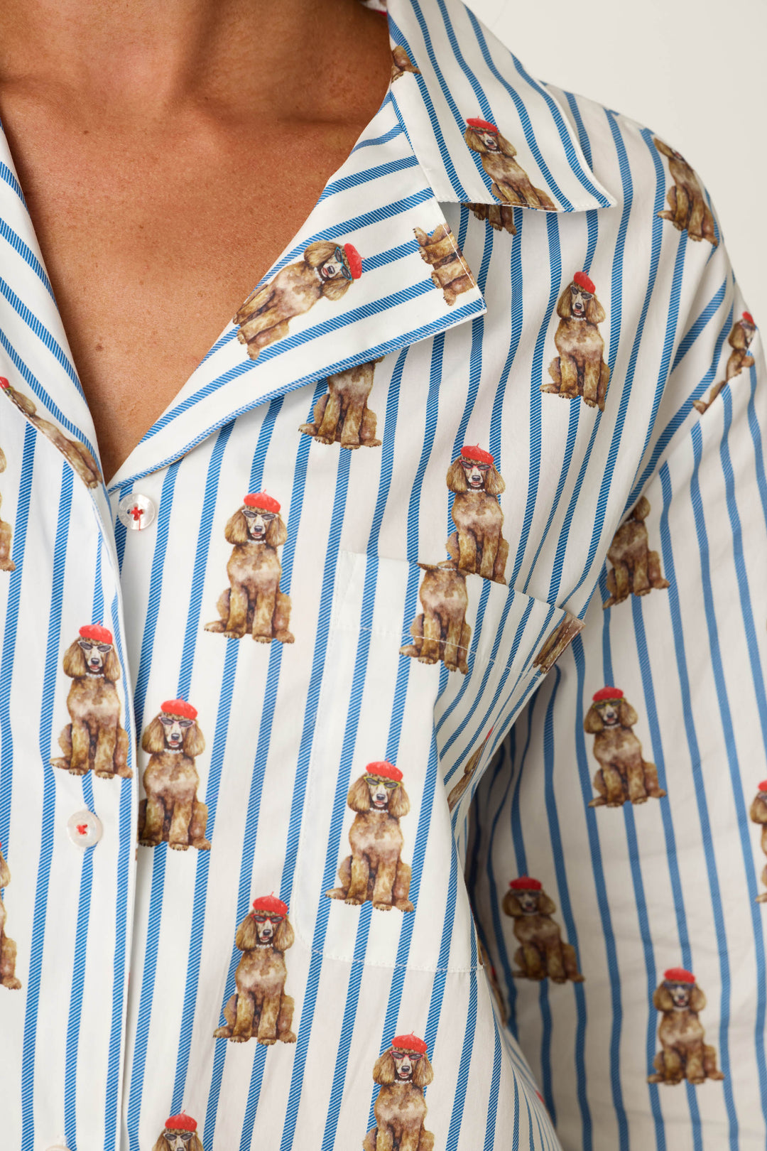 A person is shown wearing the Afterparty Poplin Stripe & Poodle Print Long Sleeve & Shorts PJ Set by P.J. Salvage, featuring blue and white stripes decorated with small brown poodles in red hats. Only the upper torso and part of the face are visible.