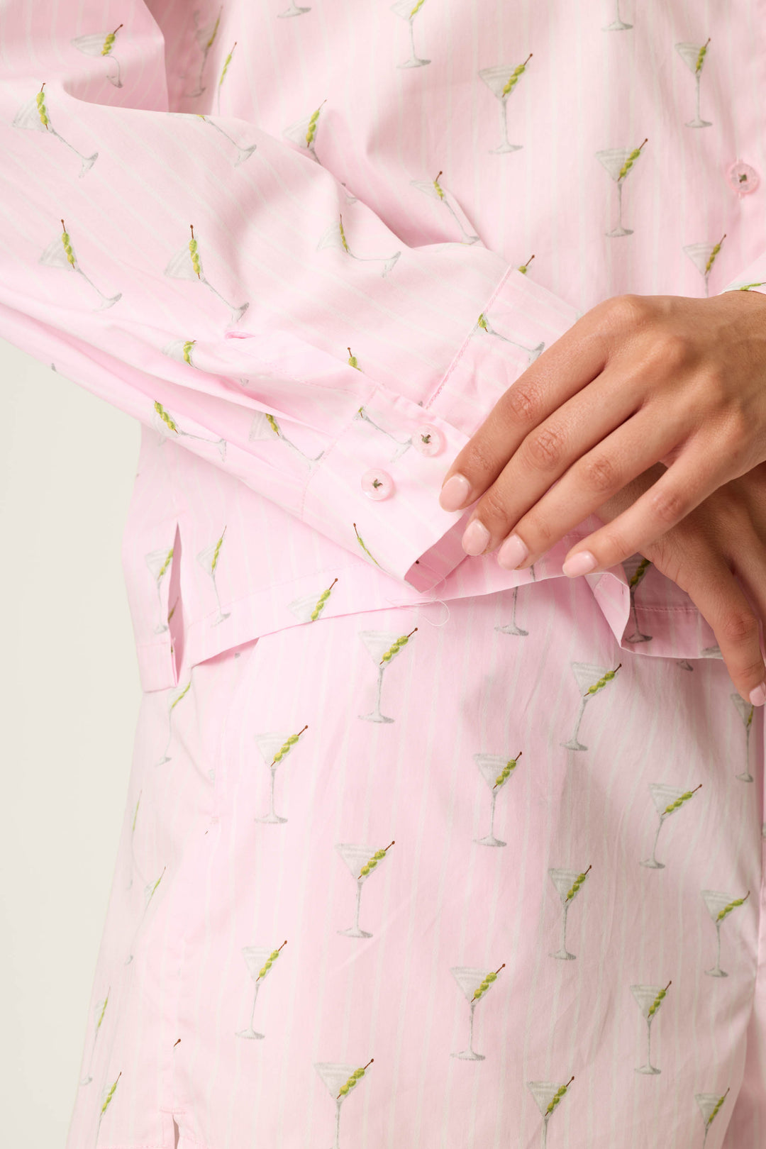 A person models the Afterparty Poplin Martini Print Long Sleeve & Shorts PJ Set by P.J. Salvage, featuring a light pink fabric decorated with a playful martini glass print. Their right hand gently rests on the buttons of the left sleeve cuff, with a plain, light-colored background behind them.