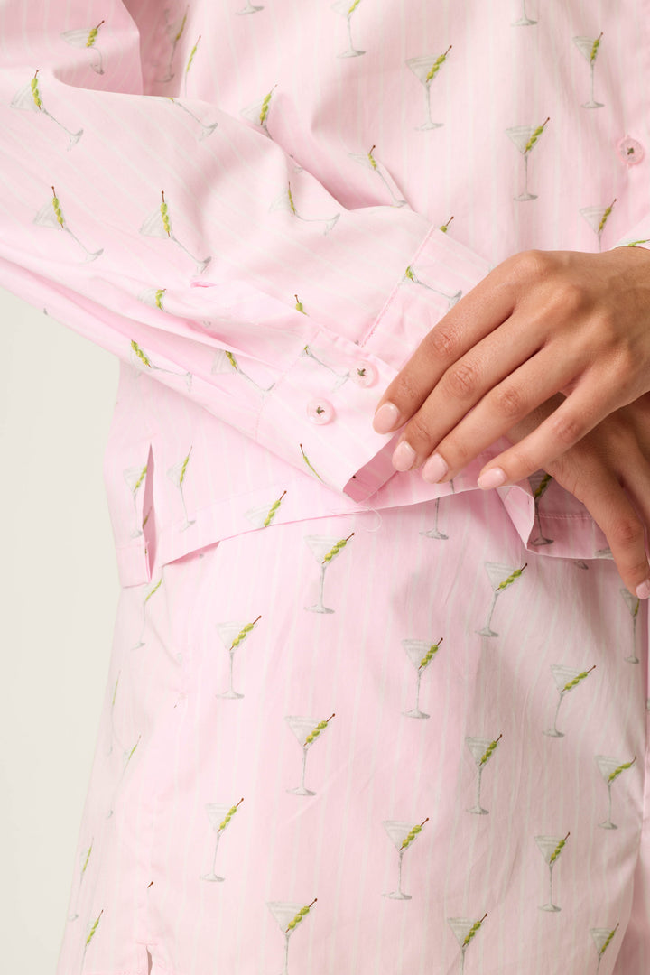 A person models the Afterparty Poplin Martini Print Long Sleeve & Shorts PJ Set by P.J. Salvage, featuring a light pink fabric decorated with a playful martini glass print. Their right hand gently rests on the buttons of the left sleeve cuff, with a plain, light-colored background behind them.
