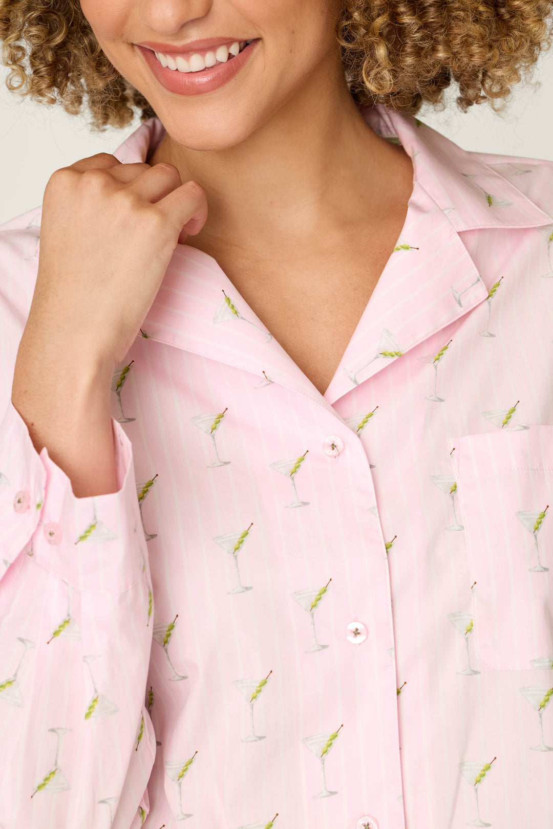 A woman with curly hair smiles while wearing the P.J. Salvage Afterparty Poplin Martini Print Long Sleeve & Shorts PJ Set, which features pink stripes and small martini glass prints. She is holding the collar with one hand.