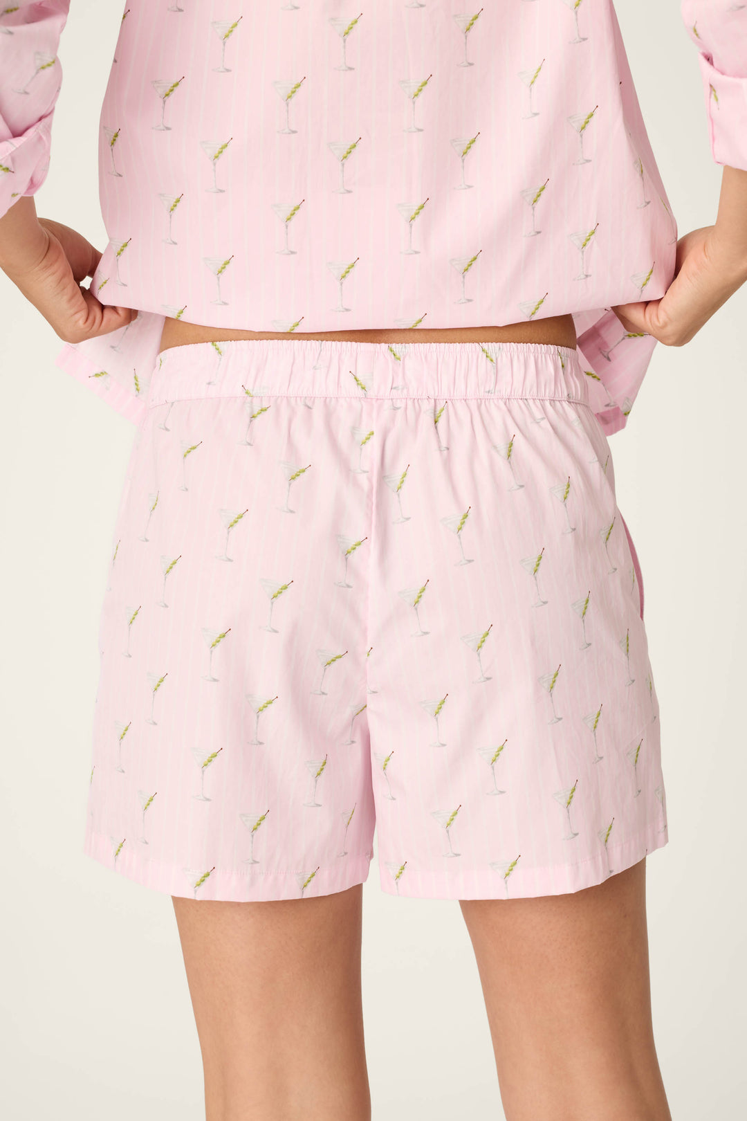 A person is shown from the back, waist to mid-thigh, wearing the Afterparty Poplin Stripe & Martini Print Pajama Shorts and matching top by P.J. Salvage, featuring a light pink color with a small martini glass pattern.