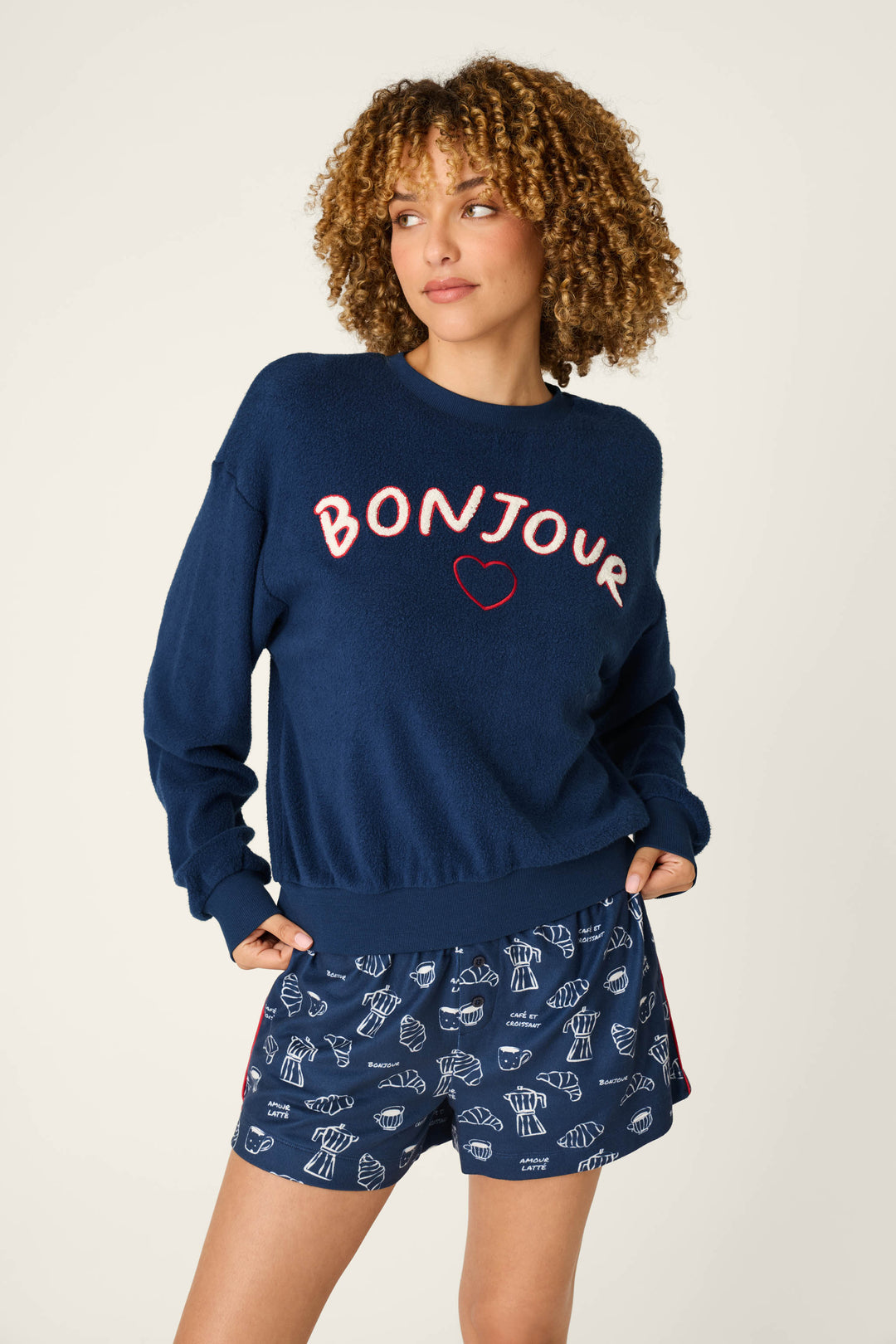A woman with curly hair stands with her hands in her pockets, gazing to the side against a light background, wearing the P.J. Salvage Bonjour Babe Boucle Bonjour Sweater—a navy top featuring "BONJOUR" and a heart—paired with matching navy shorts adorned with white croissant prints.