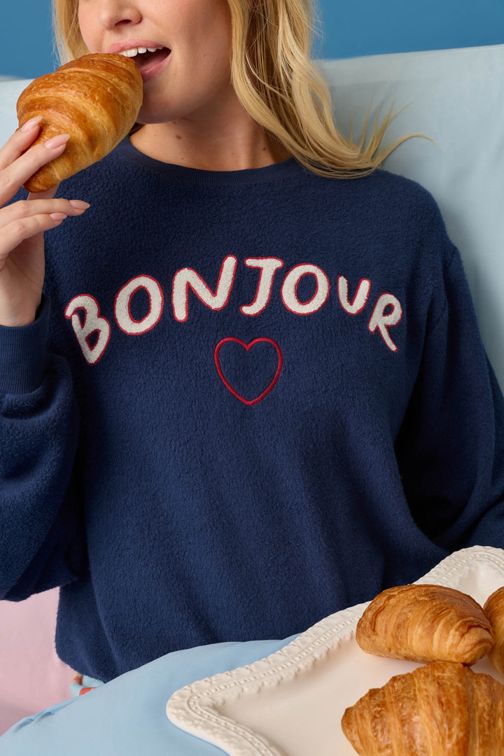 A woman in a navy blue P.J. Salvage Bonjour Babe Boucle Bonjour Sweater holds a croissant near her mouth. A plate with more croissants sits in front of her against a light blue background.