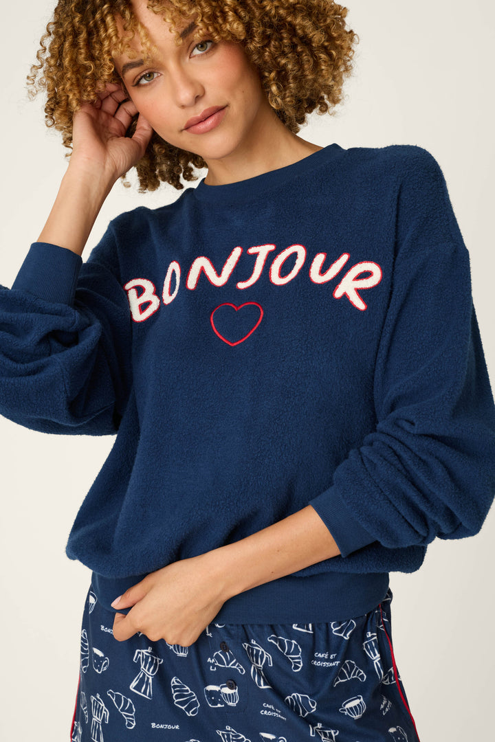 A woman with curly hair wears the P.J. Salvage Bonjour Babe Boucle Bonjour Sweater— a navy blue fuzzy sweatshirt featuring "BONJOUR" and a red heart—paired with matching pants decorated with croissant and coffee cup patterns.