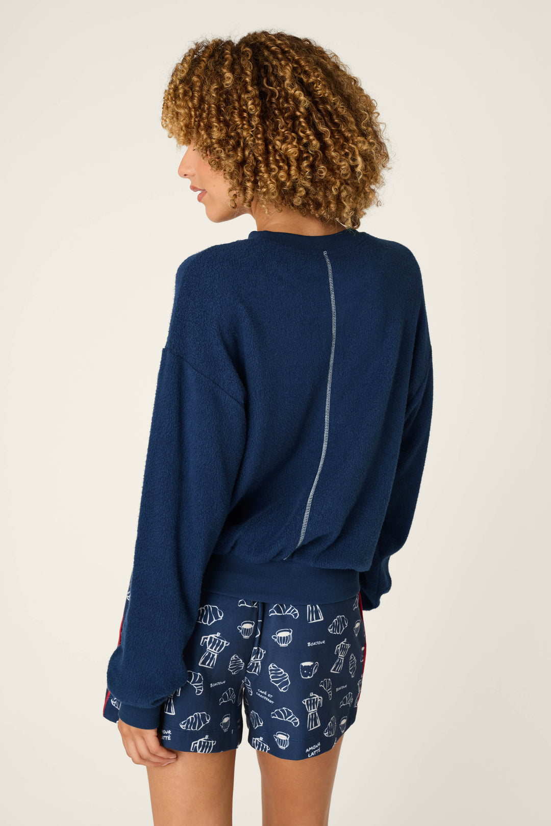 A person with curly hair stands with their back to the camera against a plain background, wearing the P.J. Salvage Bonjour Babe Boucle Bonjour Sweater in navy and matching navy shorts with white food illustrations.