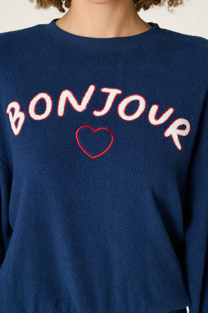 A person is shown from the neck to the upper torso wearing the P.J. Salvage Bonjour Babe Boucle Bonjour Sweater, a blue sweater featuring "BONJOUR" in white and red letters across the chest with a red heart outline below the text.
